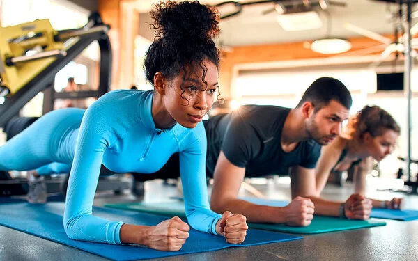 Drei Personen, eine Frau, ein Mann und eine Frau, machen Plank-Übungen auf Sportmatten in einem Fitnessstudio.