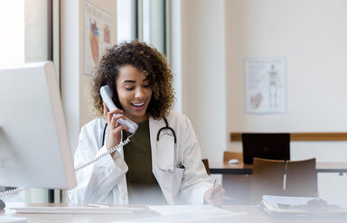 Eine junge Ärztin sitzt an ihrem Schreibtisch und telefoniert. Dabei macht sie sich Notizen auf einem Zettel.