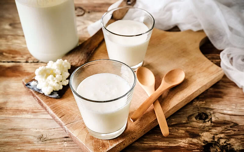 Zwei Gläser mit Kefir sowie ein Holzlöffel mit Kefirkörnern befinden sich auf einem Holzbrett, das wiederum auf einem Holztisch steht.