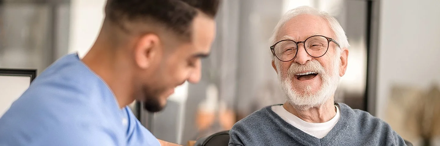Ein älterer Mann sitzt in einem Wohnzimmer auf einem Stuhl. Neben ihm sitzt ein Pfleger, der dem älteren Mann mit einem Igel-Ball die rechte Handinnenfläche mobilisiert. Beide lachen dabei.