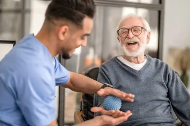 Ein älterer Mann sitzt in einem Wohnzimmer auf einem Stuhl. Neben ihm sitzt ein Pfleger, der dem älteren Mann mit einem Igel-Ball die rechte Handinnenfläche mobilisiert. Beide lachen dabei.
