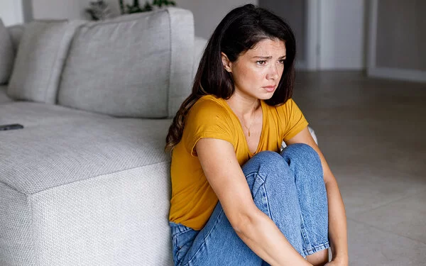 Eine Frau in einem gelben T-Shirt sitzt zusammengekauert neben einem Sofa.