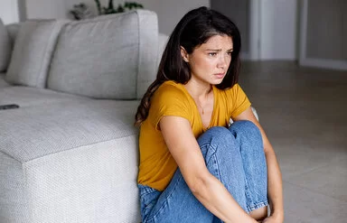 Eine Frau in einem gelben T-Shirt sitzt zusammengekauert neben einem Sofa.