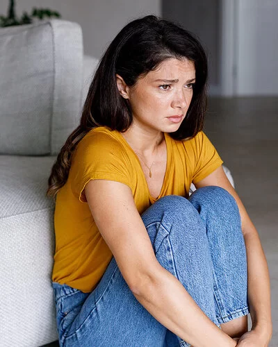Eine Frau in einem gelben T-Shirt sitzt zusammengekauert neben einem Sofa.