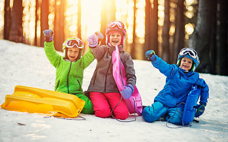 Drei Kinder sitzen mit ihren Schlitten im Schnee.