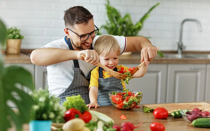 Vater und kleiner Sohn sitzen mit umgebundenen Schürzen am Küchentisch und bereiten einen bunten Salat zu.