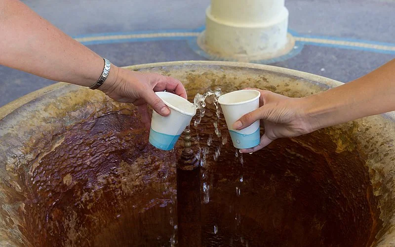 Zwei Hände fangen Wasser aus einem Quellauslauf in Pappbechern auf; innen braune Mineralspuren am Beckenrand.