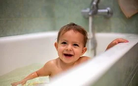 Ein lachendes Kleinkind sitzt in einer weißen Badewanne voller Wasser und hält sich mit einer Hand am Rand fest.