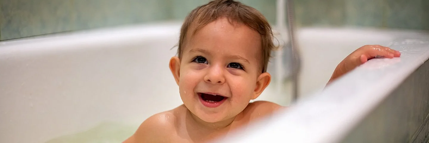 Ein lachendes Kleinkind sitzt in einer weißen Badewanne voller Wasser und hält sich mit einer Hand am Rand fest.