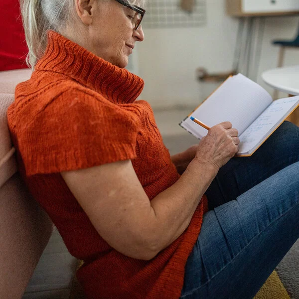 Eine ältere Frau mit dunkler Brille und orangefarbenem kurzärmeligen Rollkragenpullover, die vor einem Couchtisch am Boden sitzt, schreibt etwas in ein Notizbuch.
