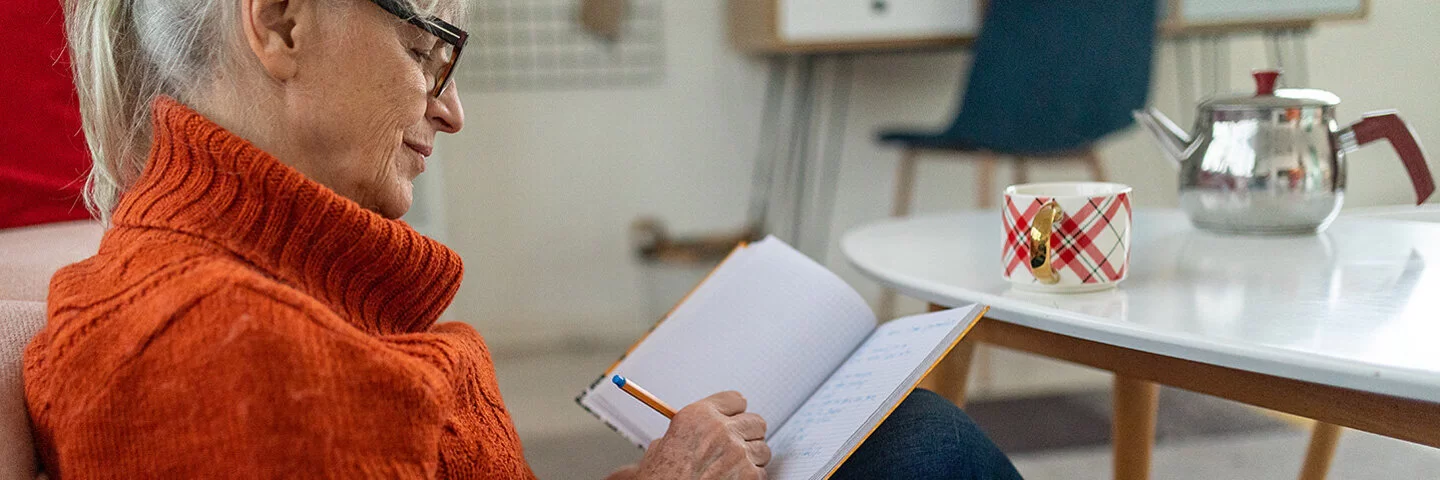 Eine ältere Frau mit dunkler Brille und orangefarbenem kurzärmeligen Rollkragenpullover, die vor einem Couchtisch am Boden sitzt, schreibt etwas in ein Notizbuch.