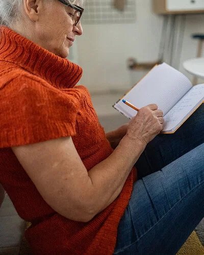 Eine ältere Frau mit dunkler Brille und orangefarbenem kurzärmeligen Rollkragenpullover, die vor einem Couchtisch am Boden sitzt, schreibt etwas in ein Notizbuch.