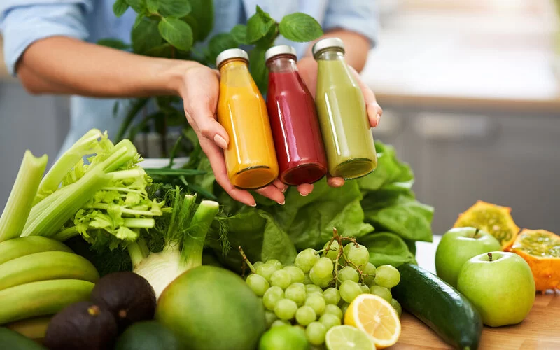 Eine junge Frau steht über einem Haufen Obst und Gemüse und hält drei kleine Flaschen mit bunten Säften in den Händen.