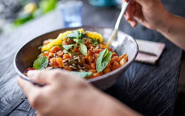 Auf einem Holztisch steht eine Schüssel mit einem Kichererbsensalat. Eine Person hält die Schüssel mit einer Hand fest und in der anderen Hand hält sie eine Gabel.