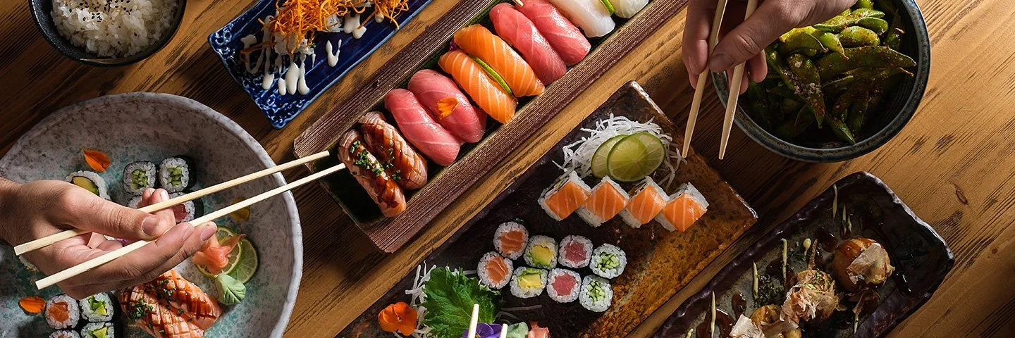 Auf einem Tisch stehen längliche Schalen mit Sushi-Varianten. Zu sehen sind auch Hände mit Stäbchen, die auf Sushi-Rollen zugreifen.