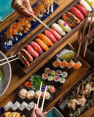 Auf einem Tisch stehen längliche Schalen mit Sushi-Varianten. Zu sehen sind auch Hände mit Stäbchen, die auf Sushi-Rollen zugreifen.