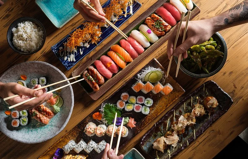 Auf einem Tisch stehen längliche Schalen mit Sushi-Varianten. Zu sehen sind auch Hände mit Stäbchen, die auf Sushi-Rollen zugreifen.