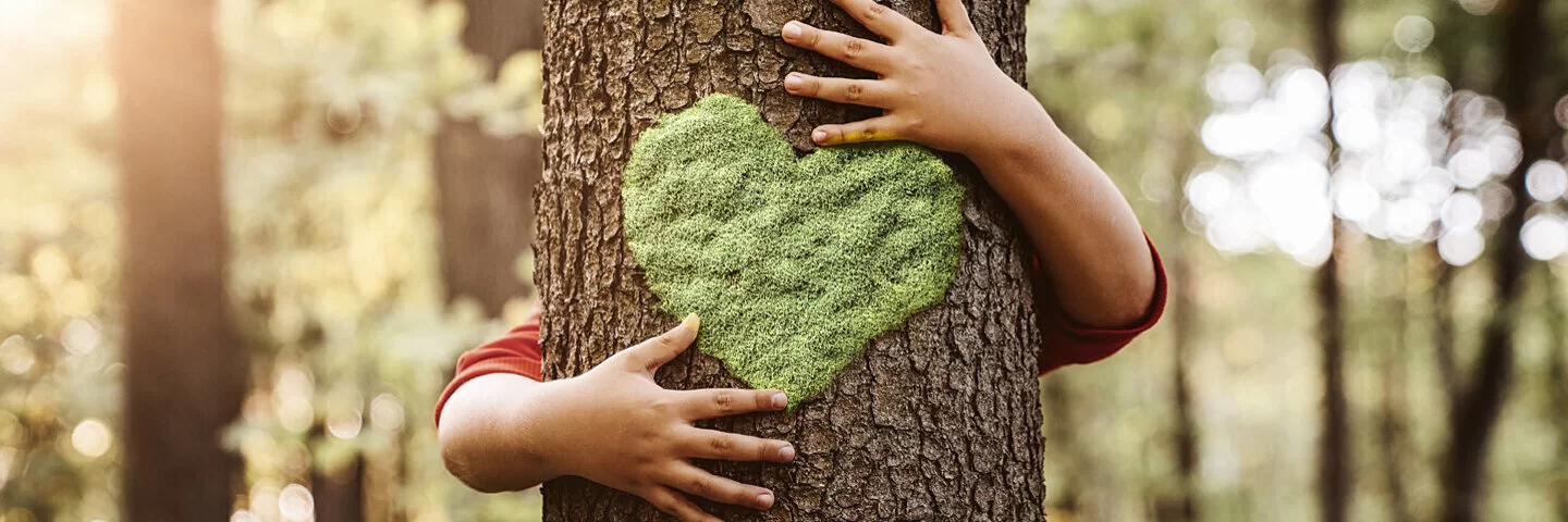 Ein Kind umarmt zum Zeichen der Nachhaltigkeit einen Baum, auf den ein grünes Herz gemalt ist.