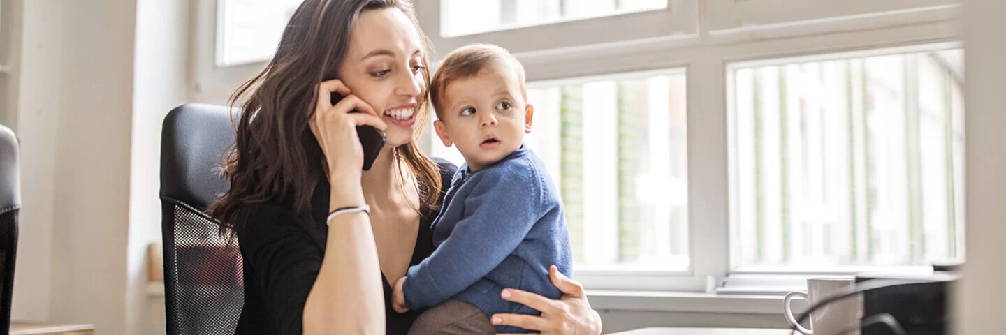 Eine junge Frau hält einen Jungen auf dem linken Arm, während sie mit der rechten Hand ein Handy ans Ohr hält und telefoniert.