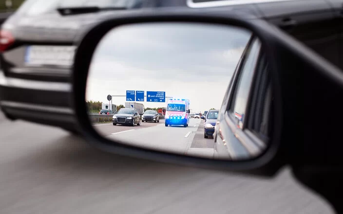 Nahaufnahme eines Auto-Seitenspiegels. Darin ist ein Krankenwagen, der von hinten näherkommt. Die Autos auf der Autobahn haben eine ordnungsgemäße Rettungsgasse gebildet.