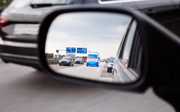 Nahaufnahme eines Auto-Seitenspiegels. Darin ist ein Krankenwagen, der von hinten näherkommt. Die Autos auf der Autobahn haben eine ordnungsgemäße Rettungsgasse gebildet.