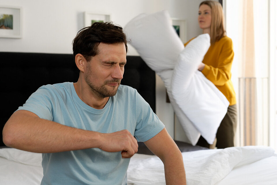 Ein Mann sitzt im Bett und muss niesen. Bei einer Hausstaubmilbenallergie der AOK in bestimmten Fällen die Kosten für spezielle Bettwäsche. 