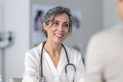 Eine nichterkennbare Frau sitzt in einer Arztpraxis, ihr gegenüber sitzt eine Ärztin und lächelt sie an.