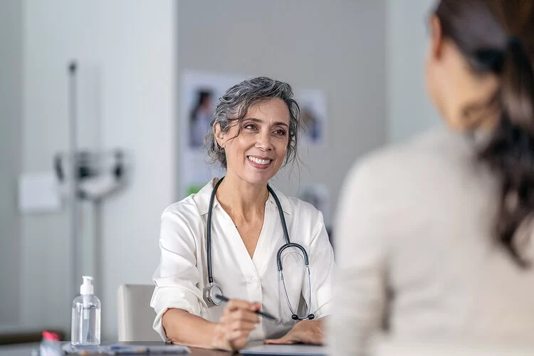 Eine nichterkennbare Frau sitzt in einer Arztpraxis, ihr gegenüber sitzt eine Ärztin und lächelt sie an.