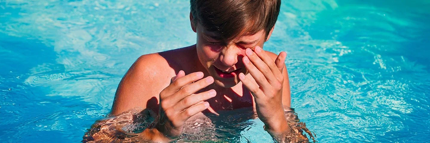 Junge im Teenager-Alter steht in einem Swimmingpool, kneift die Augen zusammen und führt die Hände zum Gesicht.