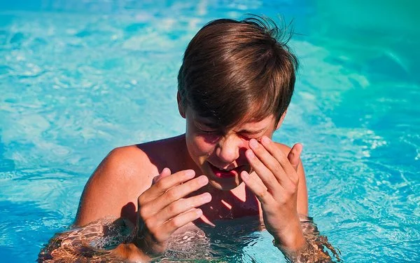 Junge im Teenager-Alter steht in einem Swimmingpool, kneift die Augen zusammen und führt die Hände zum Gesicht.