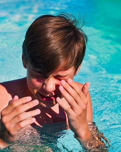 Junge im Teenager-Alter steht in einem Swimmingpool, kneift die Augen zusammen und führt die Hände zum Gesicht.