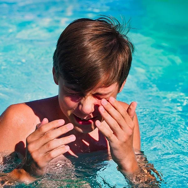Junge im Teenager-Alter steht in einem Swimmingpool, kneift die Augen zusammen und führt die Hände zum Gesicht.