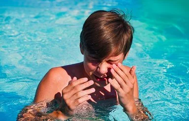 Junge im Teenager-Alter steht in einem Swimmingpool, kneift die Augen zusammen und führt die Hände zum Gesicht.