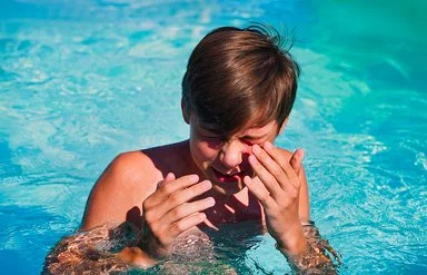 Junge im Teenager-Alter steht in einem Swimmingpool, kneift die Augen zusammen und führt die Hände zum Gesicht.