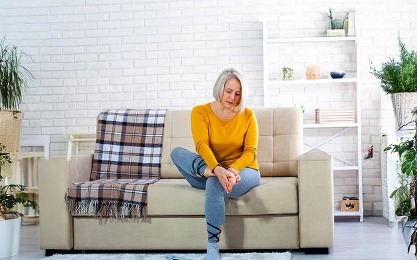 Eine Frau mittleren Alters im Wohnzimmer auf einer hellen Couch. Sie hält sich mit beiden Händen den rechten Fuß und schaut dabei besorgt.