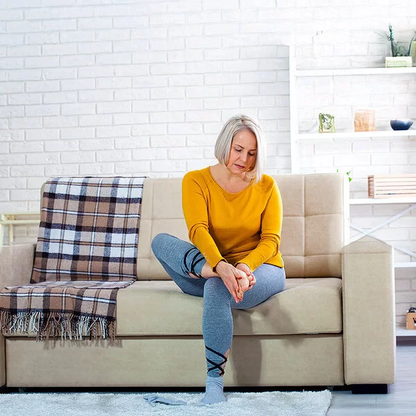Eine Frau mittleren Alters im Wohnzimmer auf einer hellen Couch. Sie hält sich mit beiden Händen den rechten Fuß und schaut dabei besorgt.