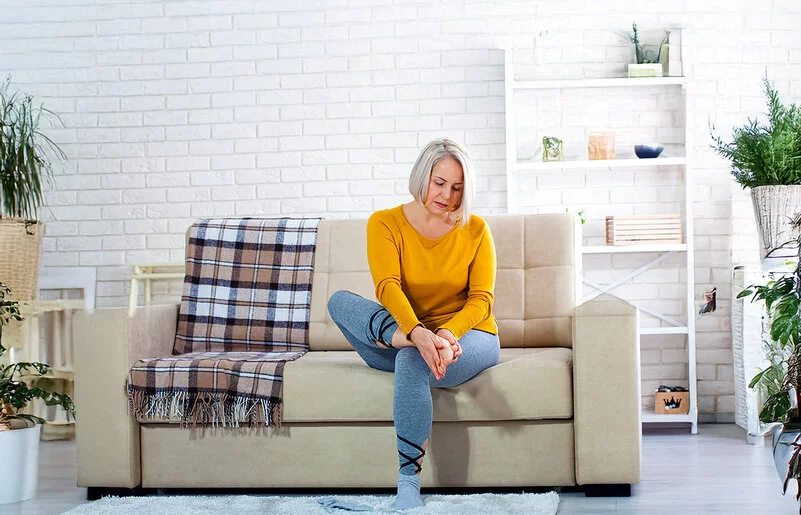 Eine Frau mittleren Alters im Wohnzimmer auf einer hellen Couch. Sie hält sich mit beiden Händen den rechten Fuß und schaut dabei besorgt.