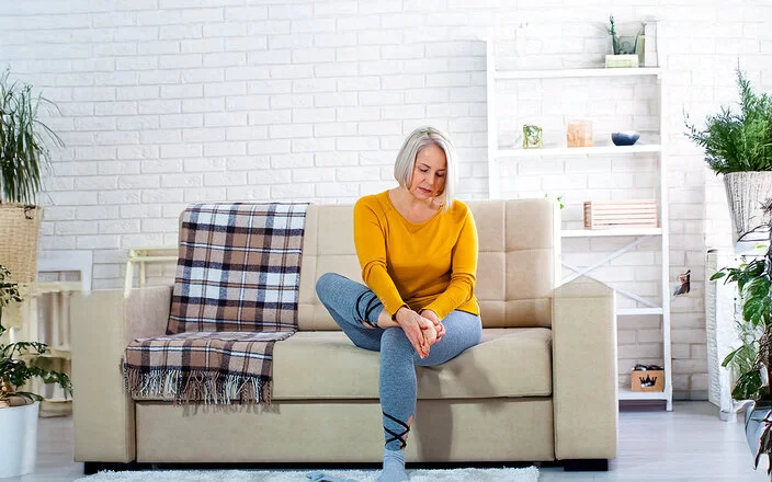 Eine Frau mittleren Alters im Wohnzimmer auf einer hellen Couch. Sie hält sich mit beiden Händen den rechten Fuß und schaut dabei besorgt.