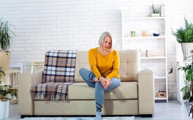 Eine Frau mittleren Alters im Wohnzimmer auf einer hellen Couch. Sie hält sich mit beiden Händen den rechten Fuß und schaut dabei besorgt.