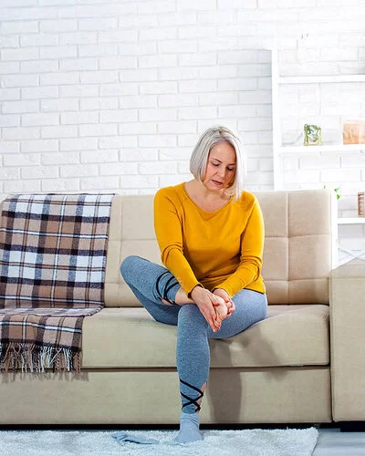 Eine Frau mittleren Alters im Wohnzimmer auf einer hellen Couch. Sie hält sich mit beiden Händen den rechten Fuß und schaut dabei besorgt.