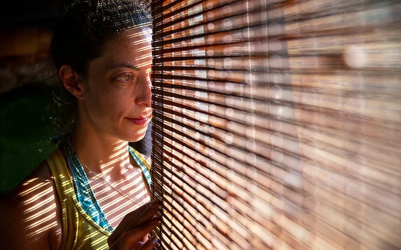 Eine Frau steht in einem abgedunkelten Raum am Fenster mit heruntergelassener Jalousie und blickt durch einen Schlitz zwischen Fenster und Jalousie nach draußen.