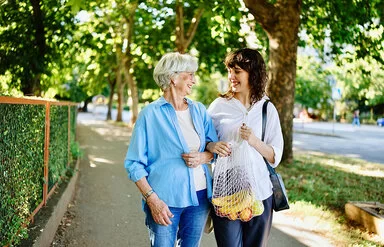 Eine junge Frau hilft einer Seniorin beim Tragen ihrer Einkäufe, beide lächeln sich an.