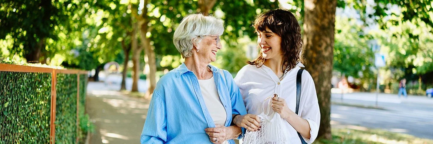 Eine junge Frau hilft einer Seniorin beim Tragen ihrer Einkäufe, beide lächeln sich an.