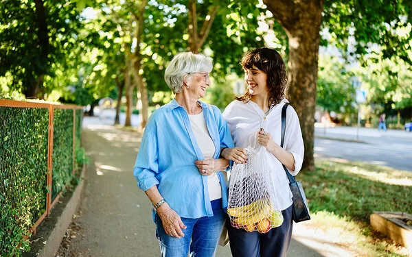 Eine junge Frau hilft einer Seniorin beim Tragen ihrer Einkäufe, beide lächeln sich an.