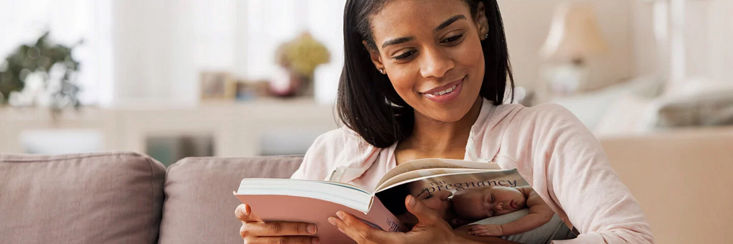 Schwangere Frau sitzt auf ihrem Sofa und liest Bücher zur Geburtsvorbereitung.