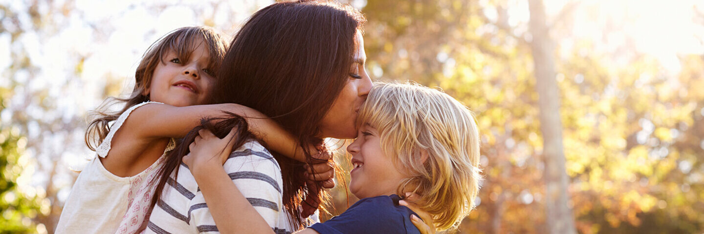 Eine Mutter mit spielt mit ihren Kindern im Park – die kleine Tochter trägt sie huckepack, den Jungen hebt sie hoch und küsst seine Stirn.