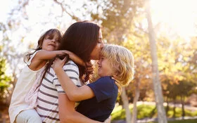 Eine Mutter mit spielt mit ihren Kindern im Park – die kleine Tochter trägt sie huckepack, den Jungen hebt sie hoch und küsst seine Stirn.