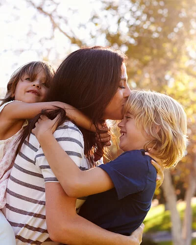 Eine Mutter mit spielt mit ihren Kindern im Park – die kleine Tochter trägt sie huckepack, den Jungen hebt sie hoch und küsst seine Stirn.