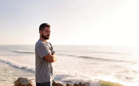 Ein Mann steht mit verschränkten Armen am Strand und schaut in den Sonnenuntergang.