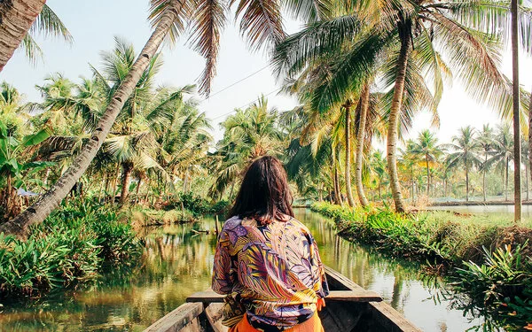 Eine Frau mit buntgemusterter Bluse sitzt in einem hölzernen Kanu und ist von hinten zu sehen. Sie fährt über einen mit Palmen gesäumten Fluss.